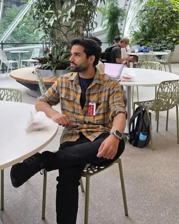 Priyanshu Yadav sitting at a table in a glass conservatory full of plants, looking to the side.
